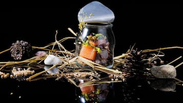A glass jar with a lid, containing a piece of salmon, radish, and some greenery, surrounded by pine cones, rocks, and straw on a reflective surface.