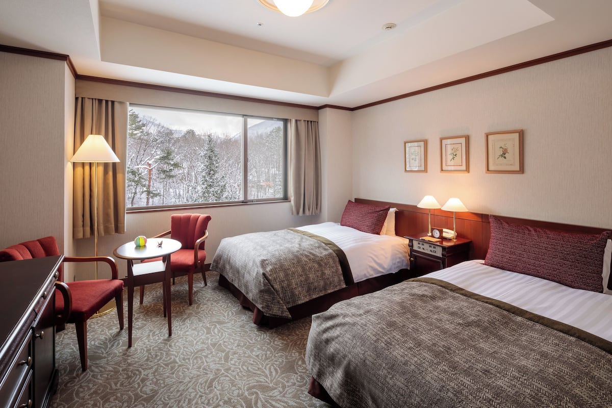 A hotel room with two beds, a desk, a chair, a table, and a window with a view of trees.