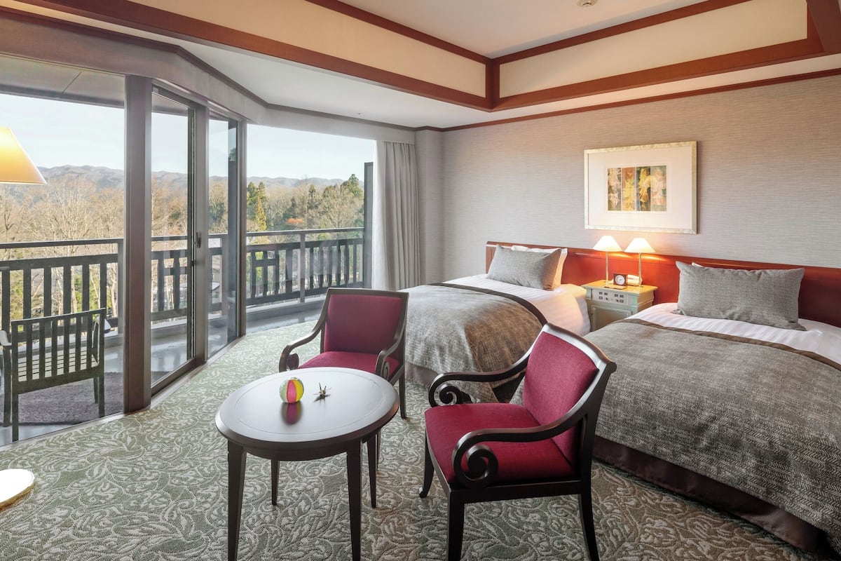 A hotel room with a bed, two chairs, a small table, and a balcony with a view of the mountains.