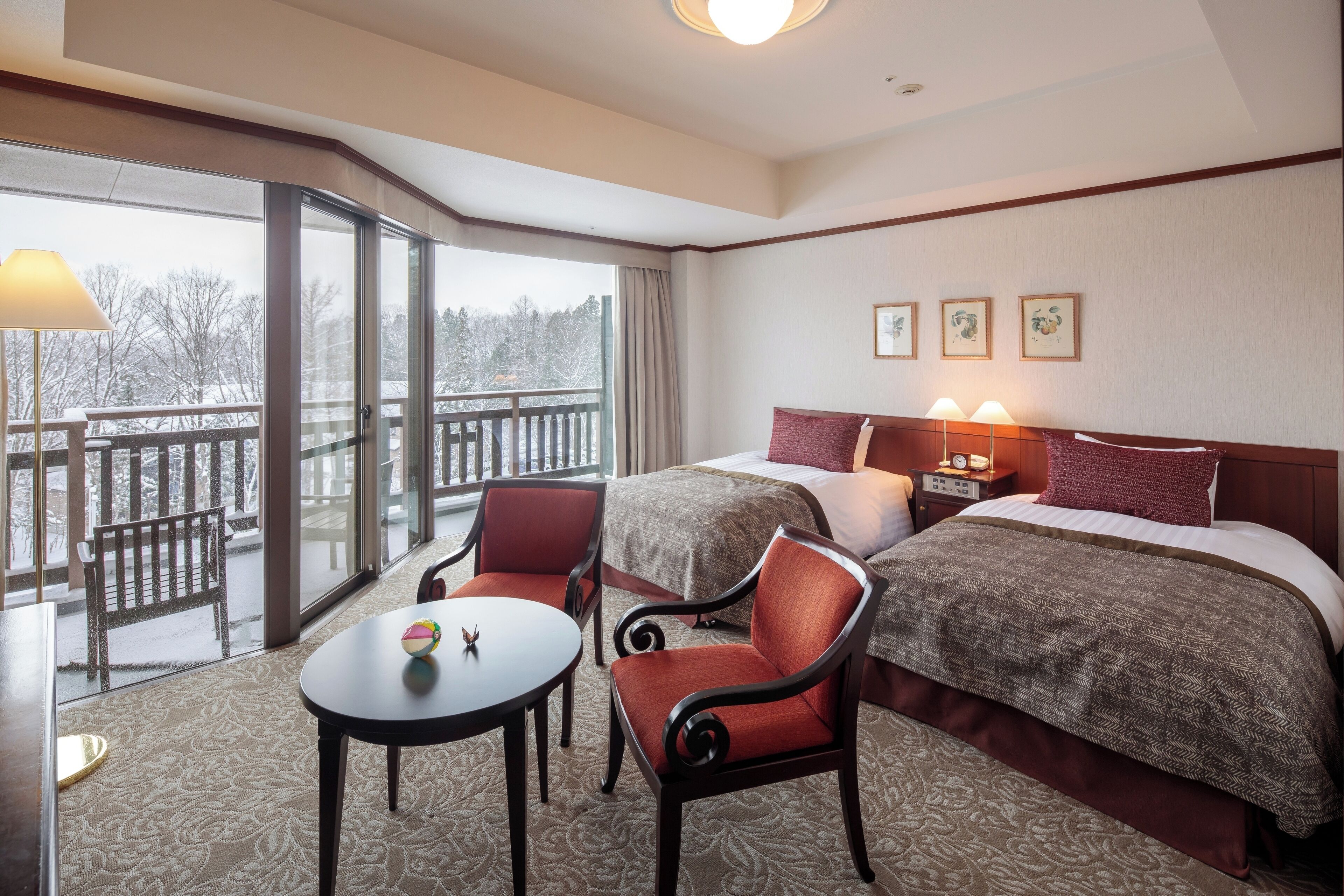A hotel room with two beds, a sitting area with a table and chairs, and a balcony with a view of trees.
