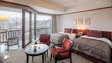 Habitación de hotel con dos camas, zona de estar con una mesa y sillas, y balcón con vistas a árboles.