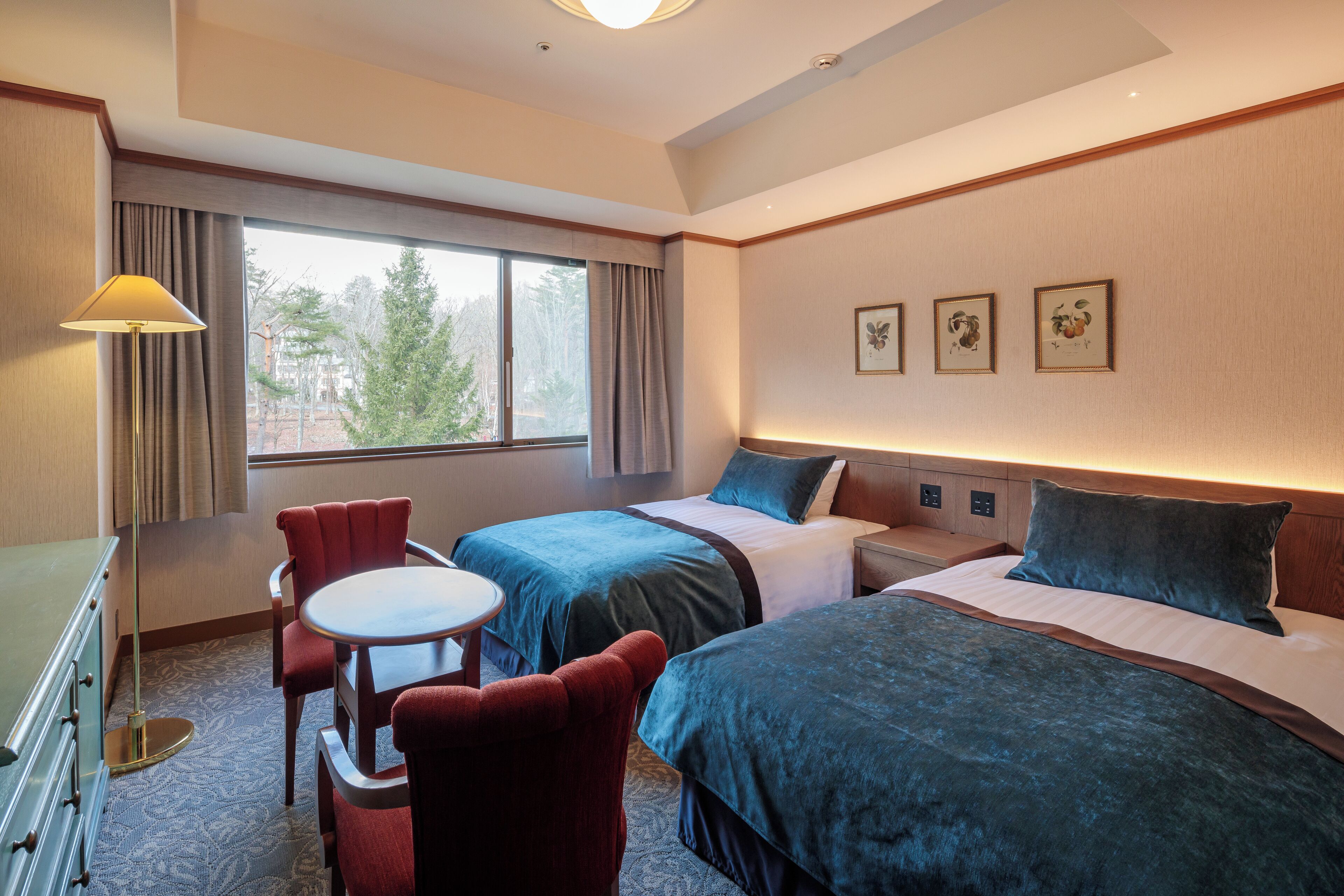 A hotel room with two beds, a desk, a chair, and a window with a view of trees.