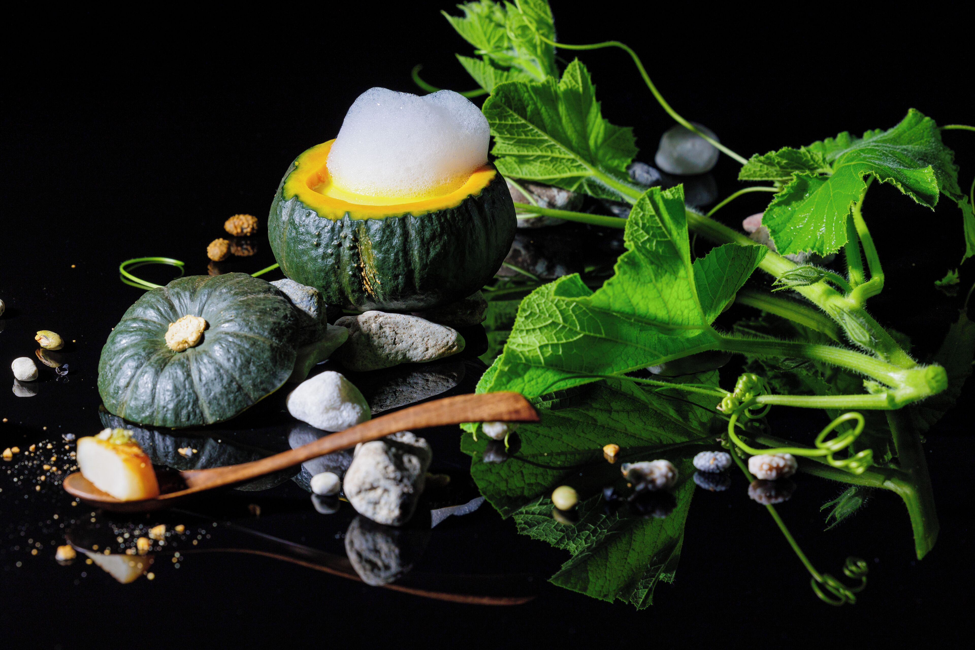 A close-up of a sliced pumpkin with a white filling, surrounded by green leaves and scattered seeds on a reflective surface.