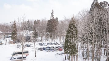 A snow-covered parking lot with several cars and a bus, surrounded by tall, leafless trees.