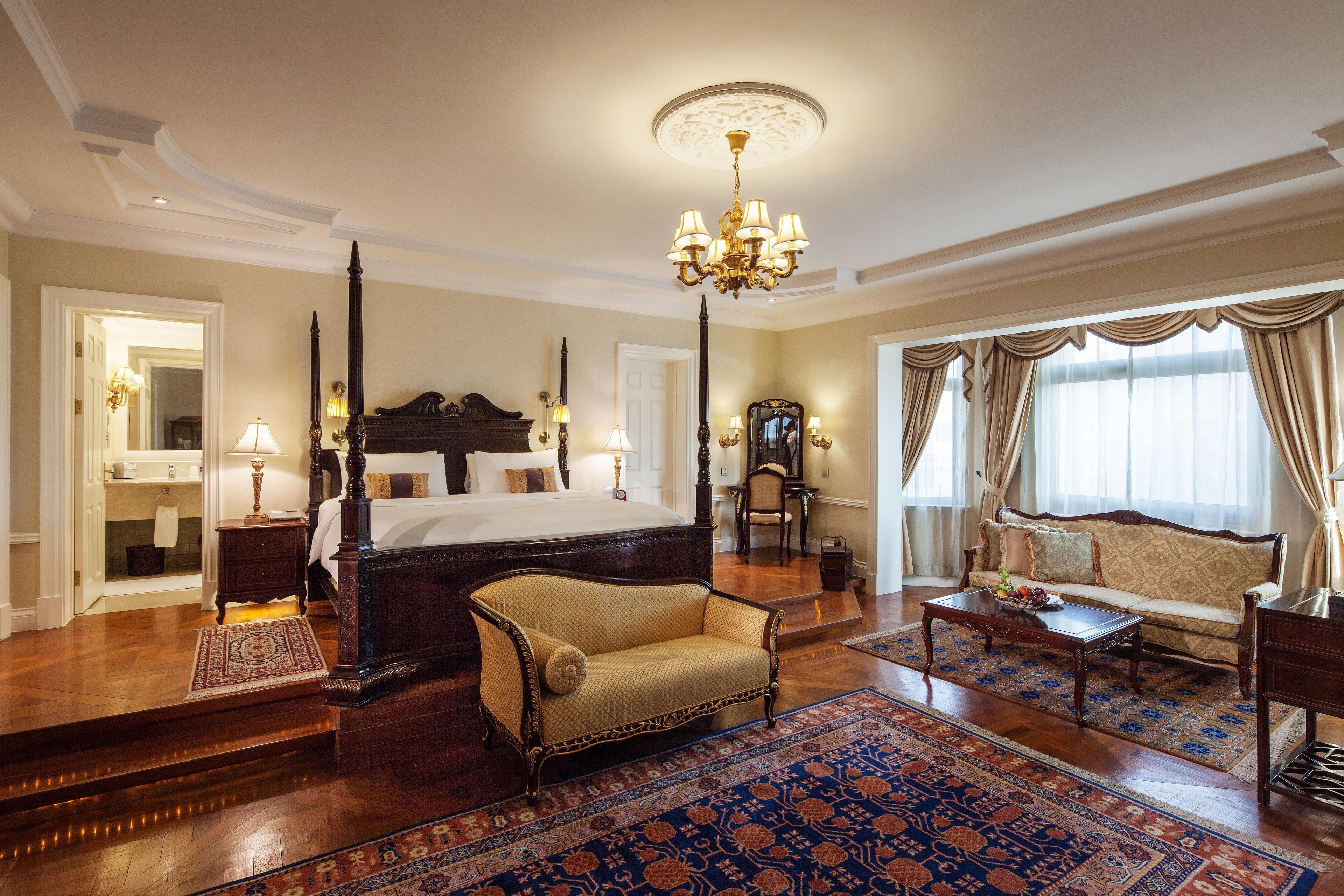 A spacious bedroom with a four-poster bed, a sitting area with a sofa, a dining table, and a chandelier.
