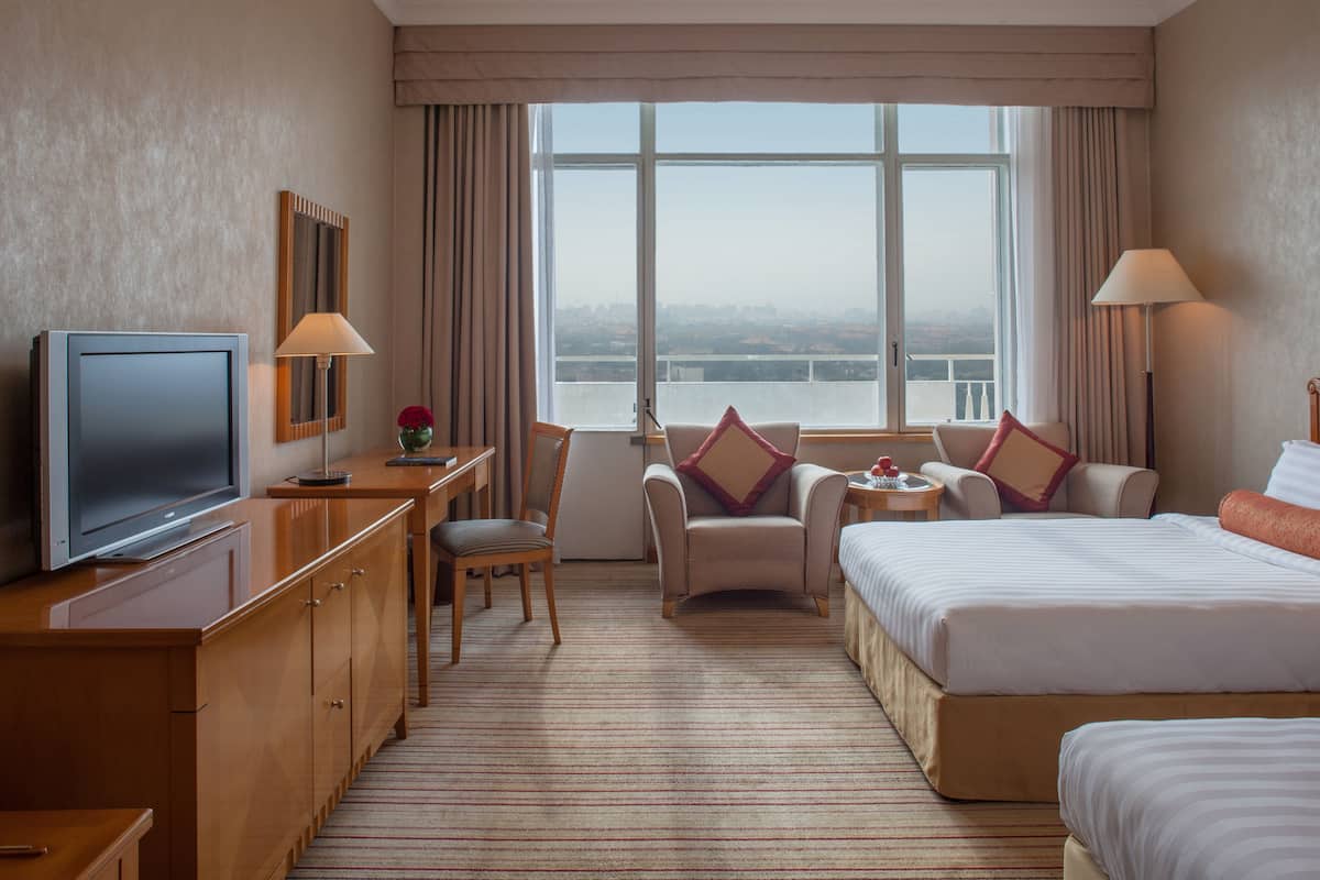 Ein Hotelzimmer mit einem großen Bett, einem Schreibtisch mit Fernseher, einem Sessel, einem kleinen Tisch mit Lampe und Blick auf die Stadt durch das Fenster.