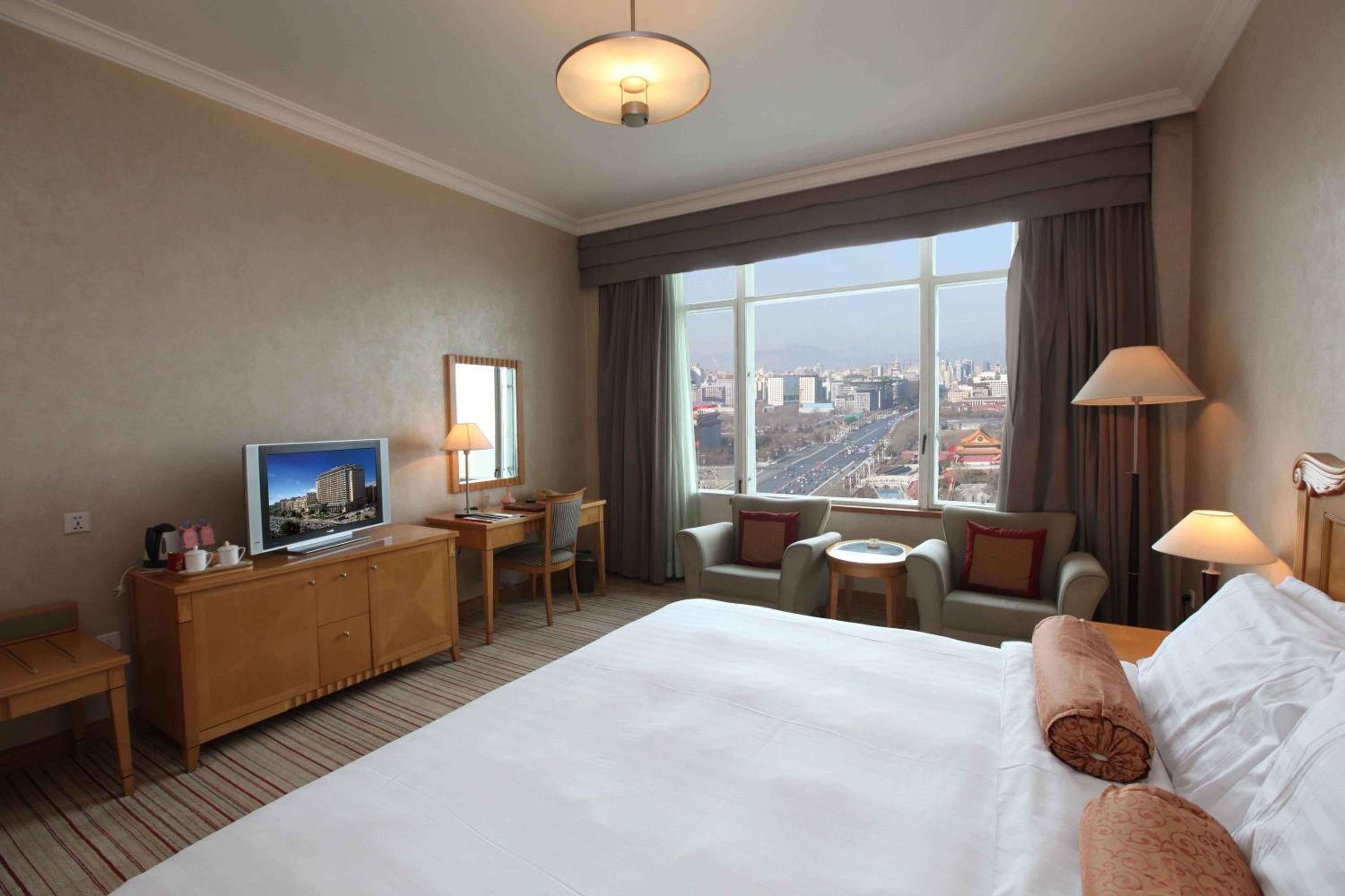 A hotel room with a large bed, a desk with a computer, a television, and a view of a cityscape through a large window.