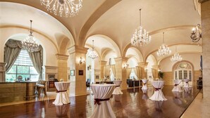 Um salão espaçoso com altos tetos arqueados e lustres, com mesas cobertas por toalhas brancas e laços vermelhos, e um espaço de bar com cadeiras e balcão.