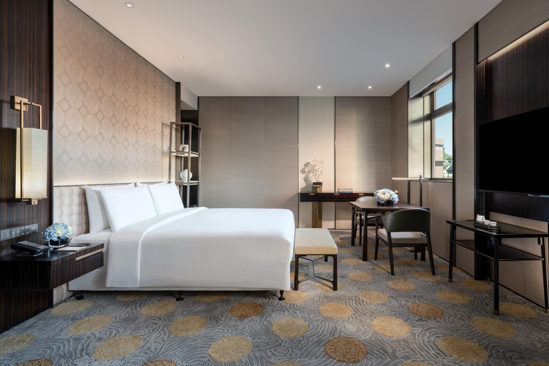 A modern hotel room with a large bed, a desk with a chair, a television, and a bookshelf.