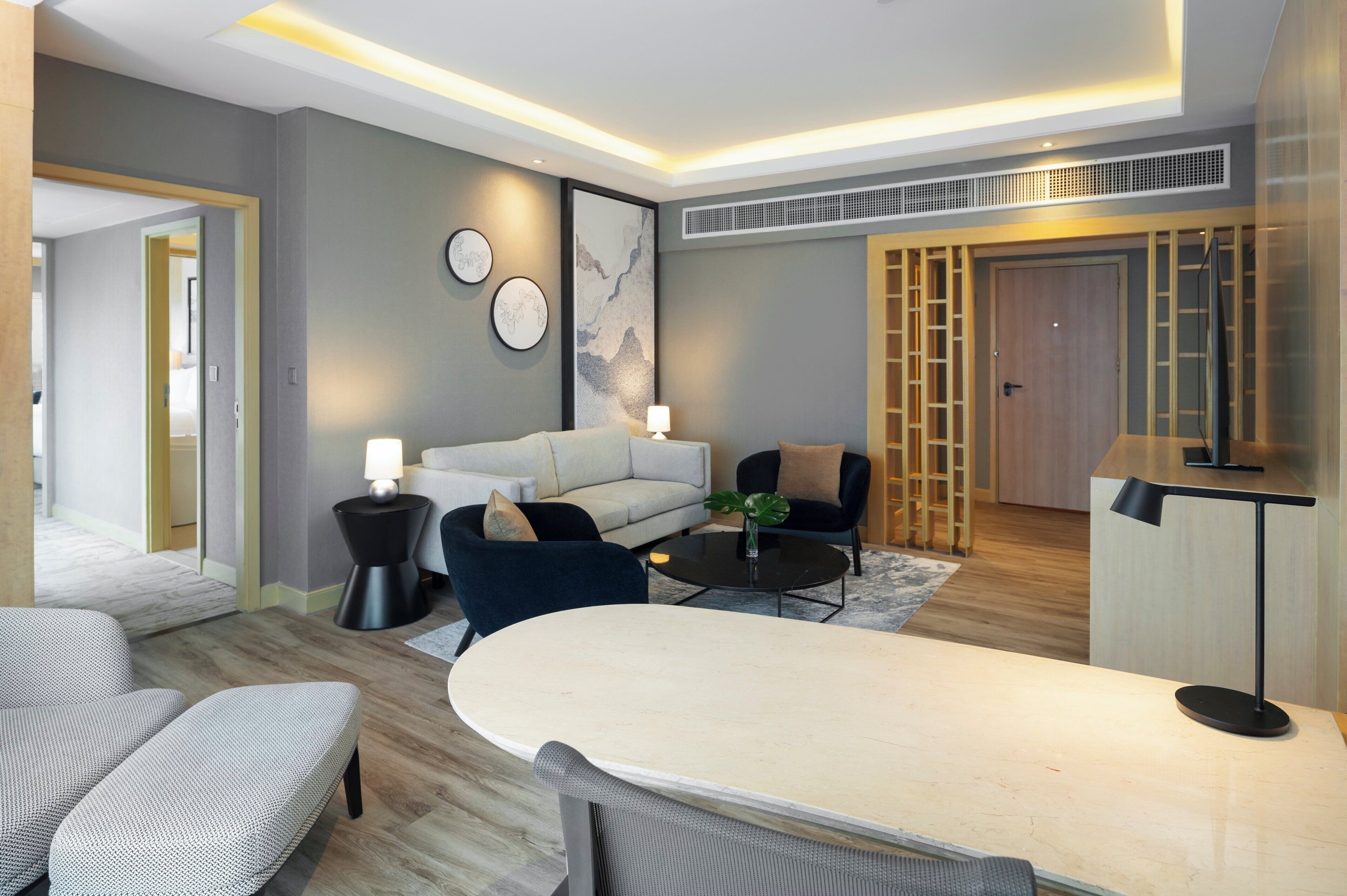 A modern hotel room with a sofa, armchairs, a dining table, and a TV.