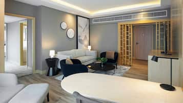 A modern hotel room with a sofa, armchairs, a dining table, and a TV.
