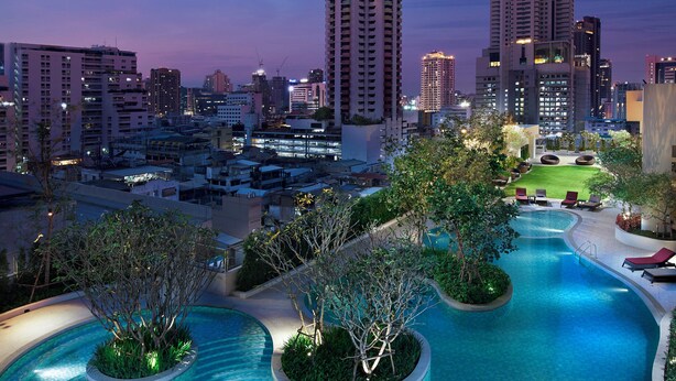 A swimming pool with a curved design, surrounded by greenery and lounge chairs, set against a cityscape with tall buildings at dusk. - Amari Bangkok (Bangkok)
