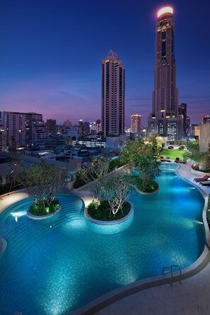 Una piscina con diseño curvo, rodeada de follaje y sillas de descanso, frente a un paisaje urbano con edificios altos al anochecer.