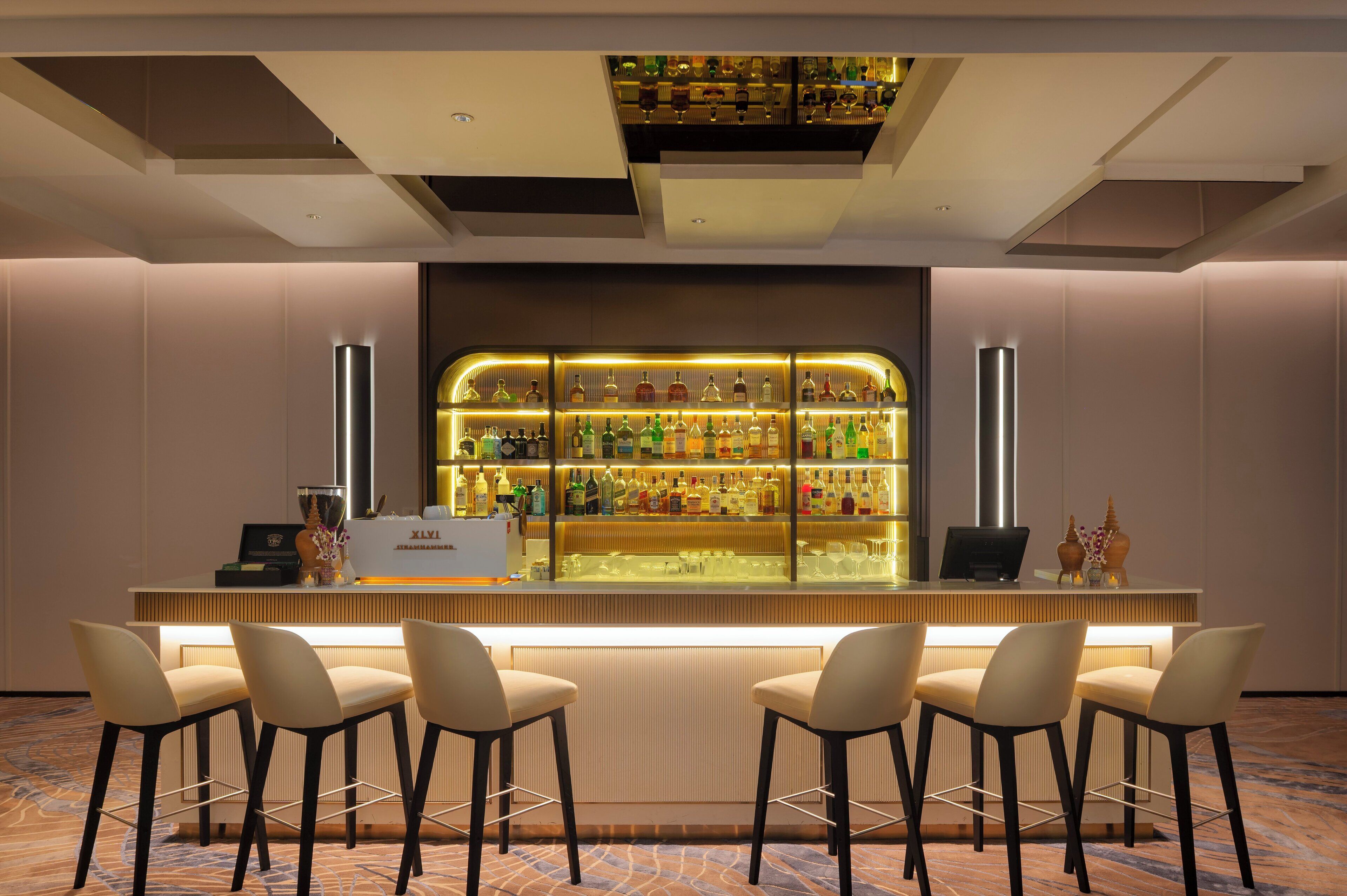 Een bar met een lange toonbank, diverse drankflessen en vier barkrukken.