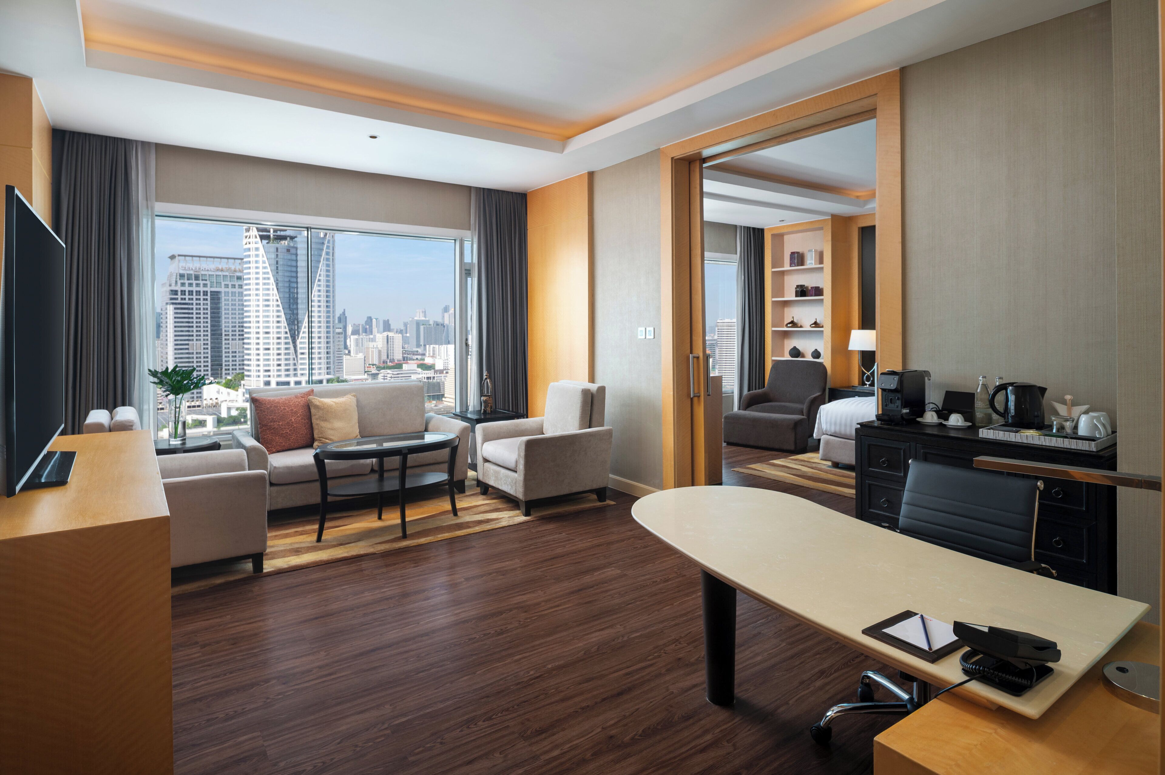 Una habitación de hotel moderna con un ventanal amplio que ofrece vistas a la ciudad, una zona de estar con sofá y sillones, una mesa de comedor y una kitchenette.