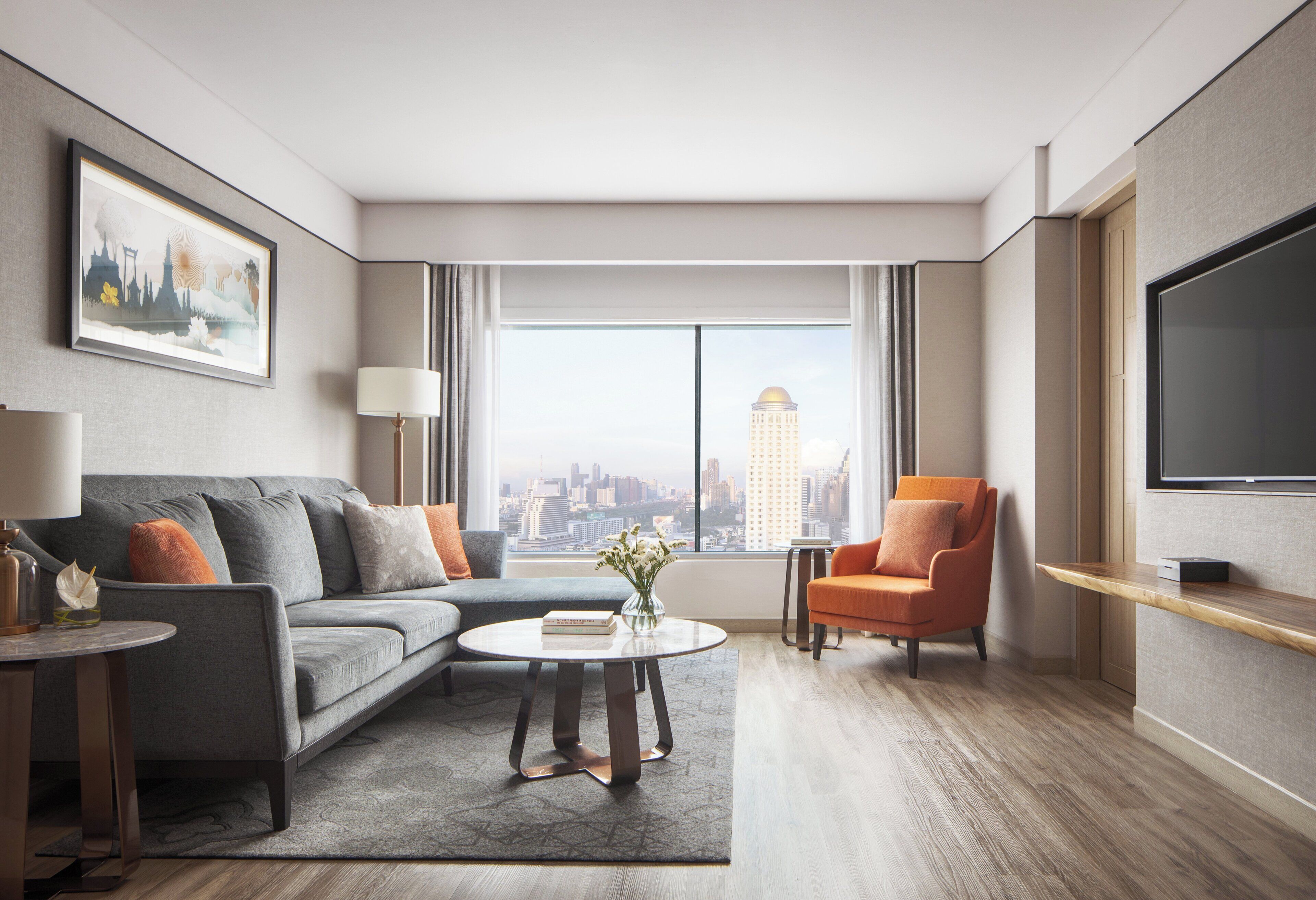 Habitación de hotel moderna con un sofá gris, una mesa de centro redonda, una mesita auxiliar, una silla, un televisor y un ventanal con vistas a la ciudad.