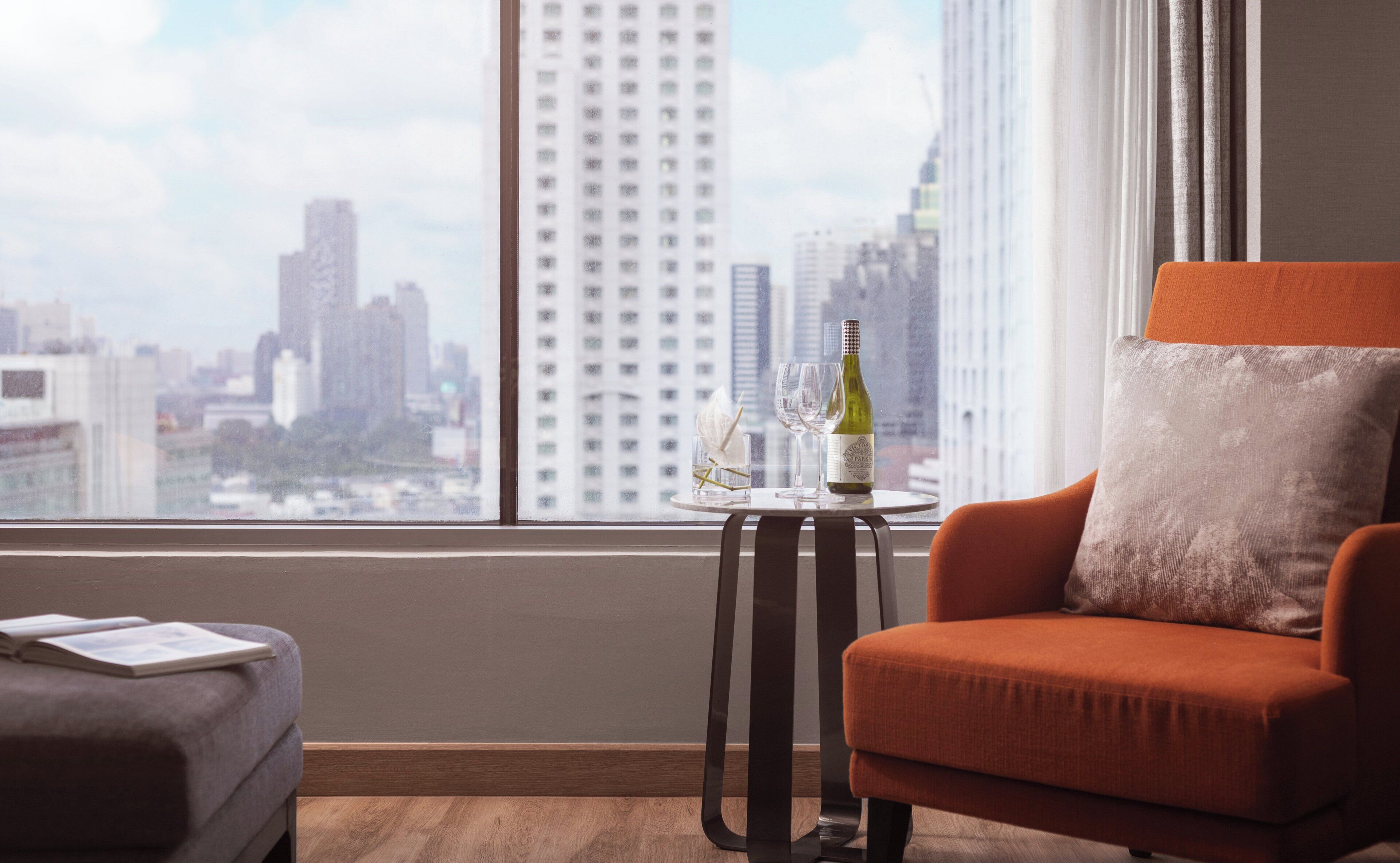 A modern room with a view of a cityscape through large windows, a small round table with a bottle and glasses, and an orange armchair.