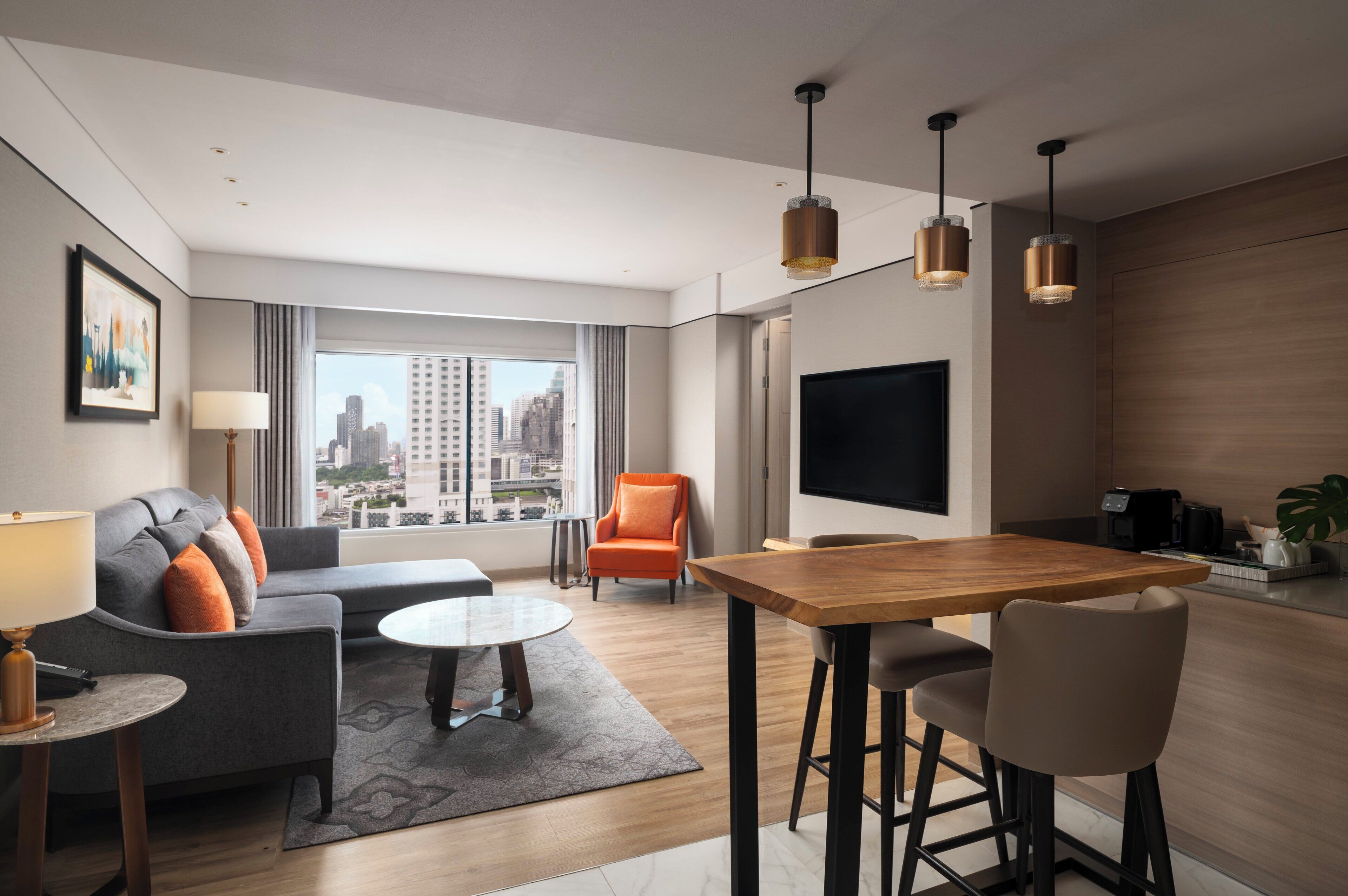 A modern hotel room with a sofa, a dining table, a TV, and a city view.
