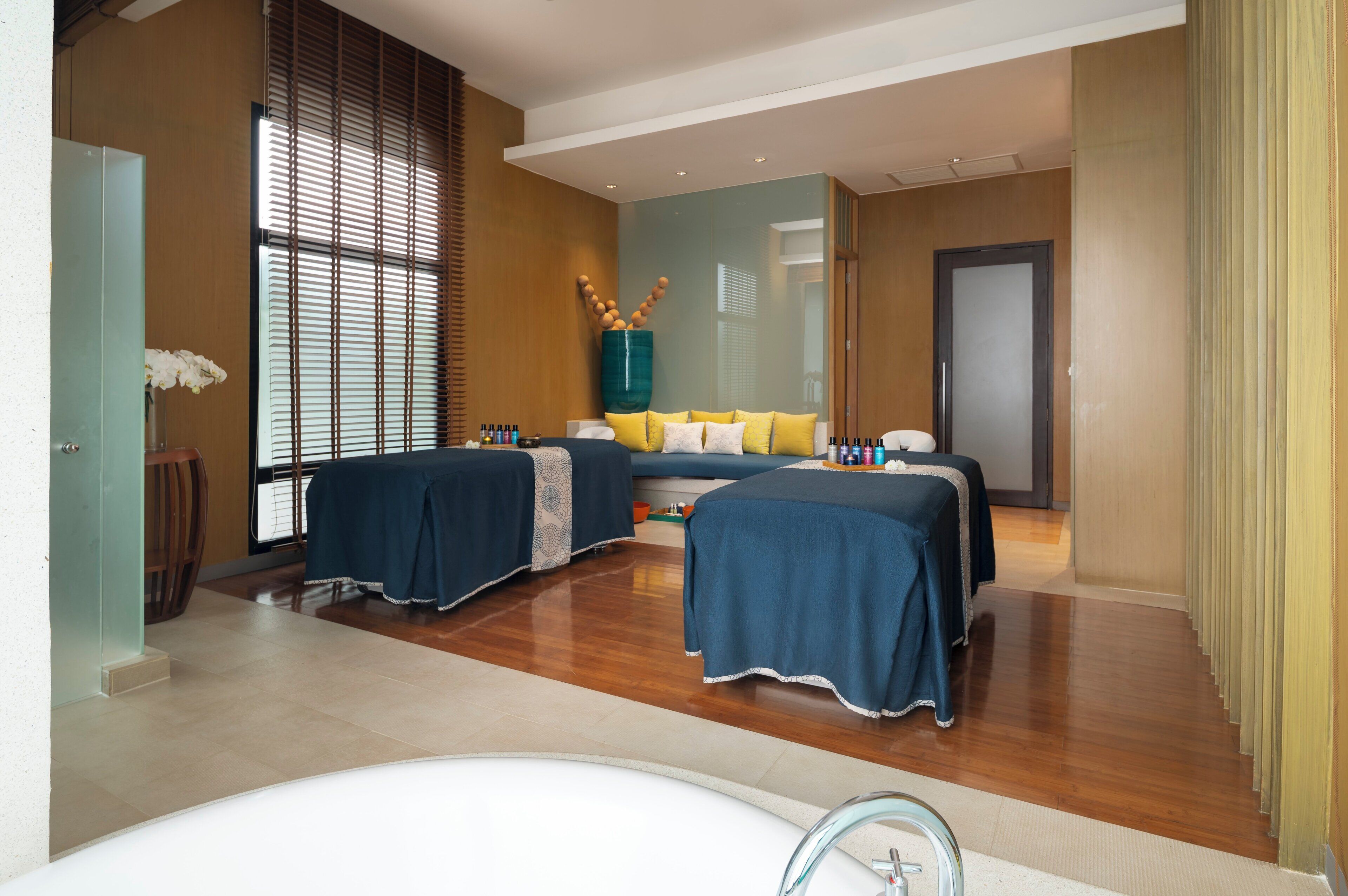 A modern hotel room with two beds, a large window with blinds, a bathtub, and a wooden floor.