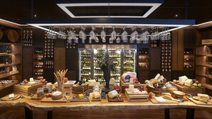 A well-lit cheese and wine tasting room with wooden shelves, a central display of cheeses and fruits, and hanging wine glasses above. - Amari Bangkok (Bangkok)
