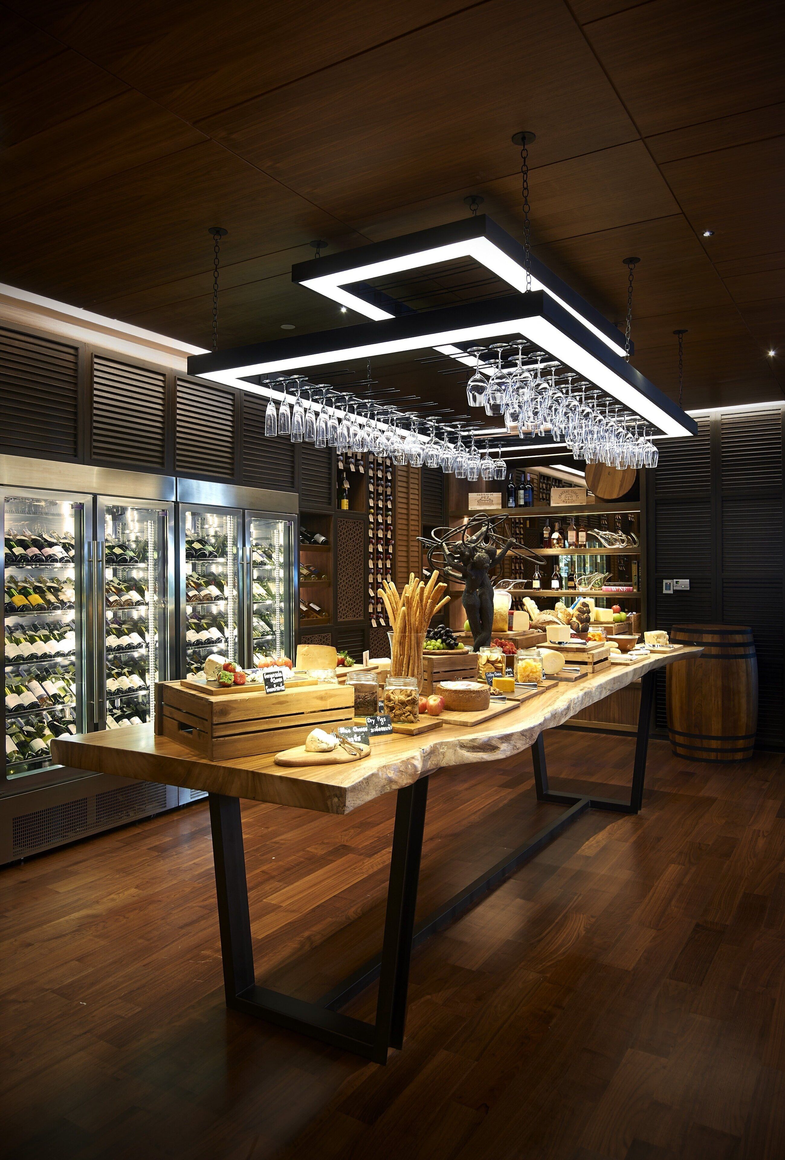 A wine tasting room with a long table displaying various cheeses and fruits, surrounded by wine coolers and shelves with bottles.