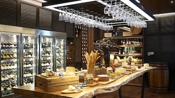 A wine tasting room with a long table displaying various cheeses and fruits, surrounded by wine coolers and shelves with bottles.