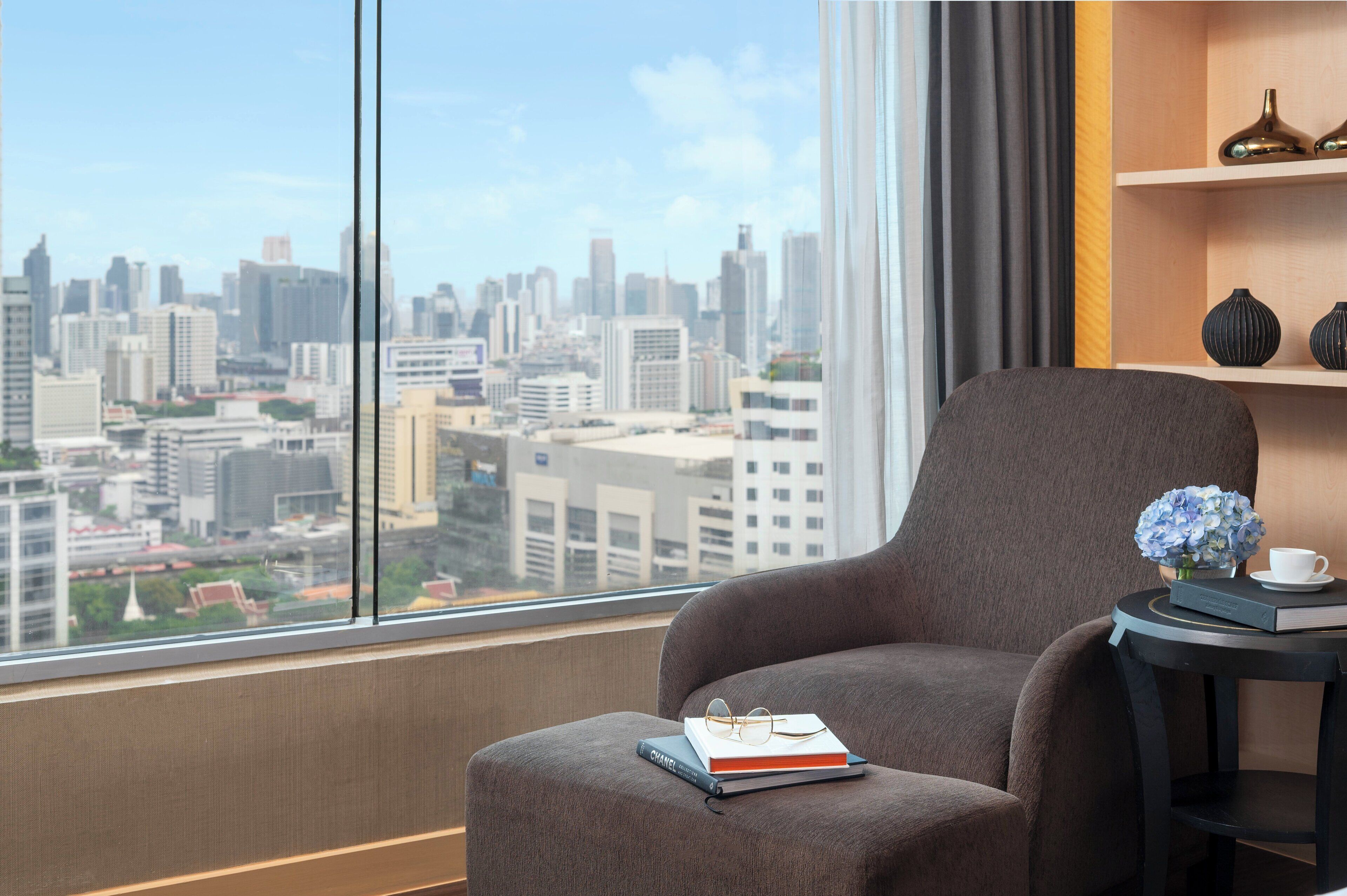 Una habitación moderna con un ventanal grande que ofrece vistas a la ciudad, un sillón cómodo, una mesita con un libro y vasos, y un jarrón decorativo con flores.