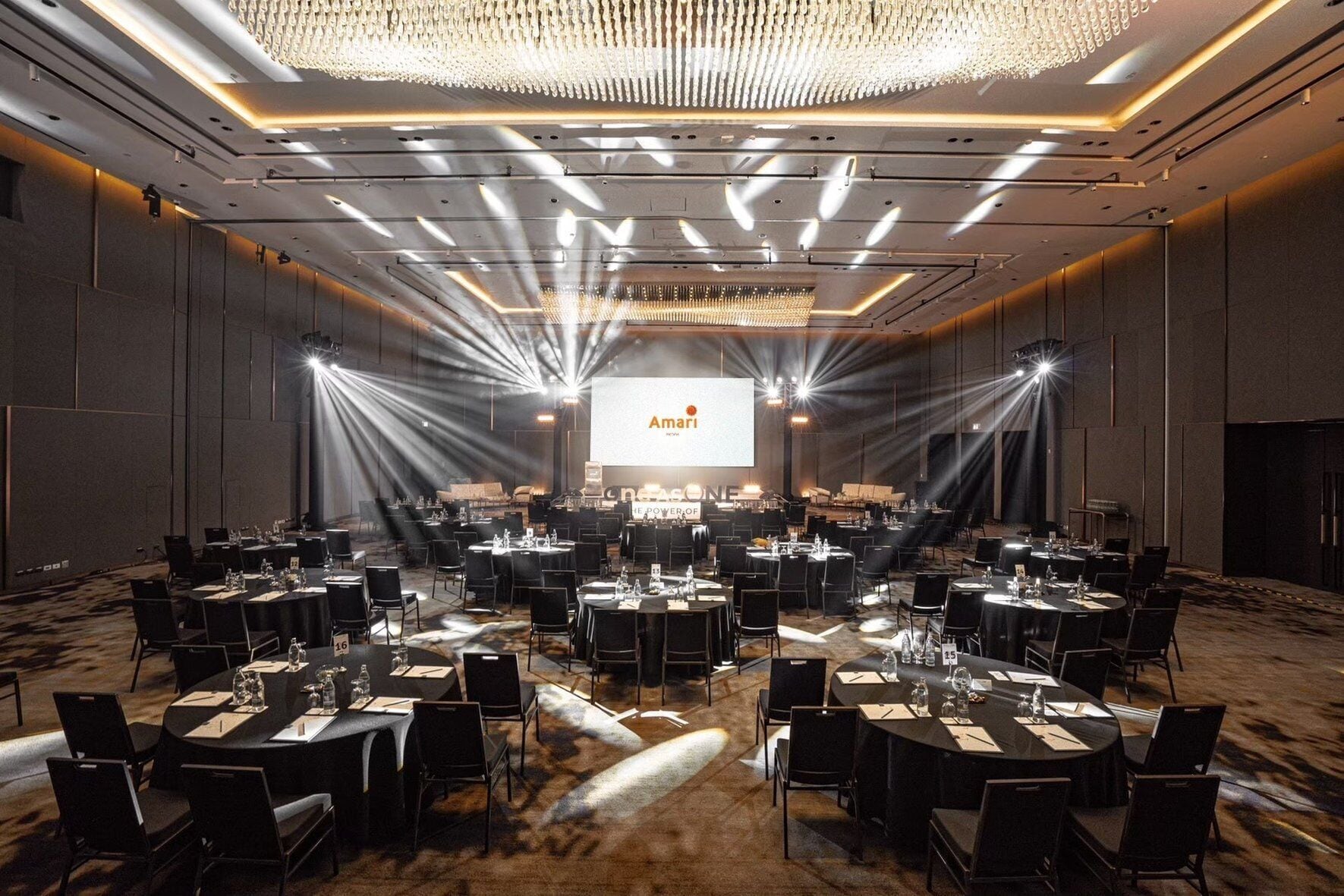 A large conference room with multiple tables and chairs, a central stage with a screen displaying 'Amari', and a chandelier.