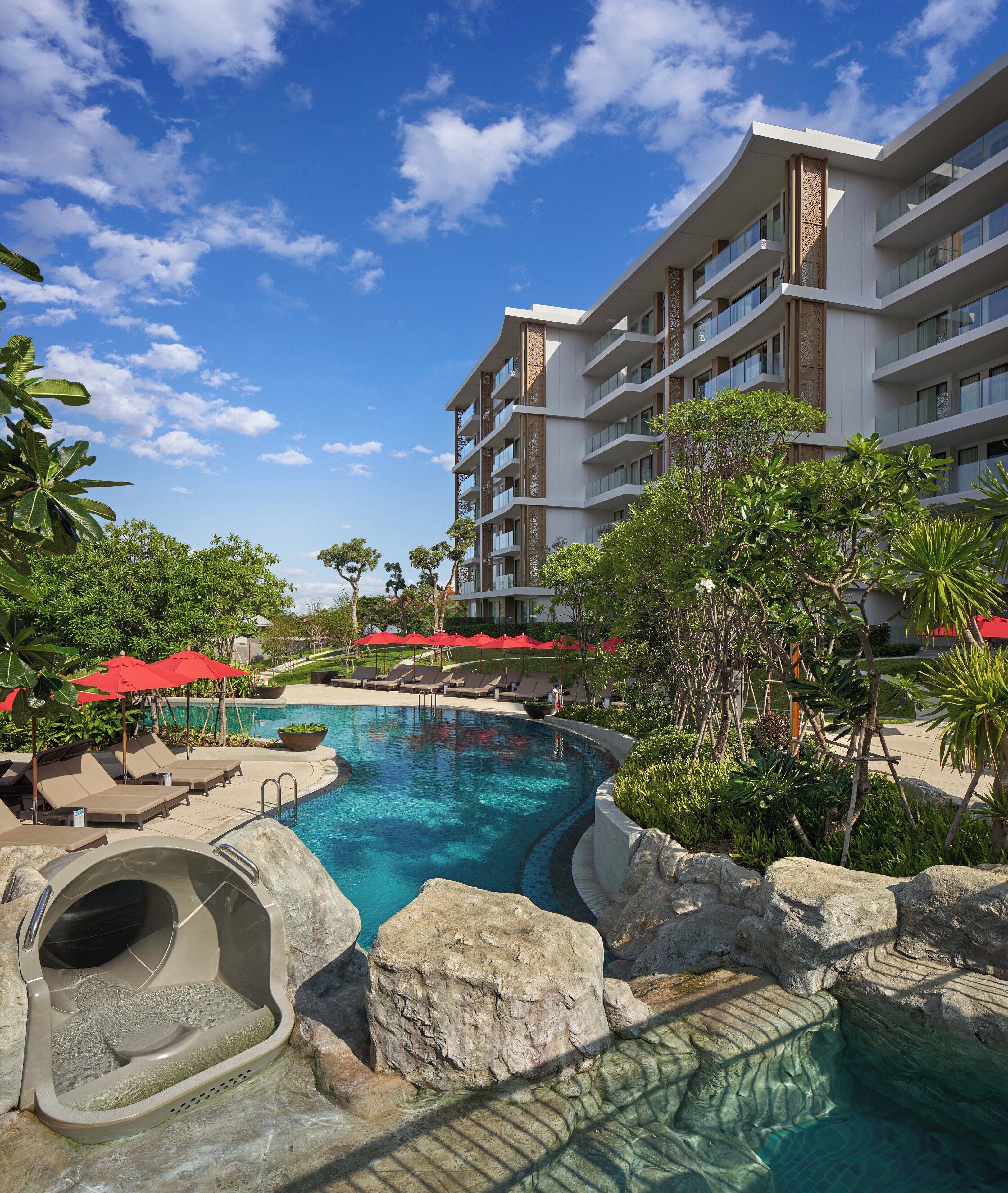 A hotel with a swimming pool, lounge chairs, and red umbrellas.