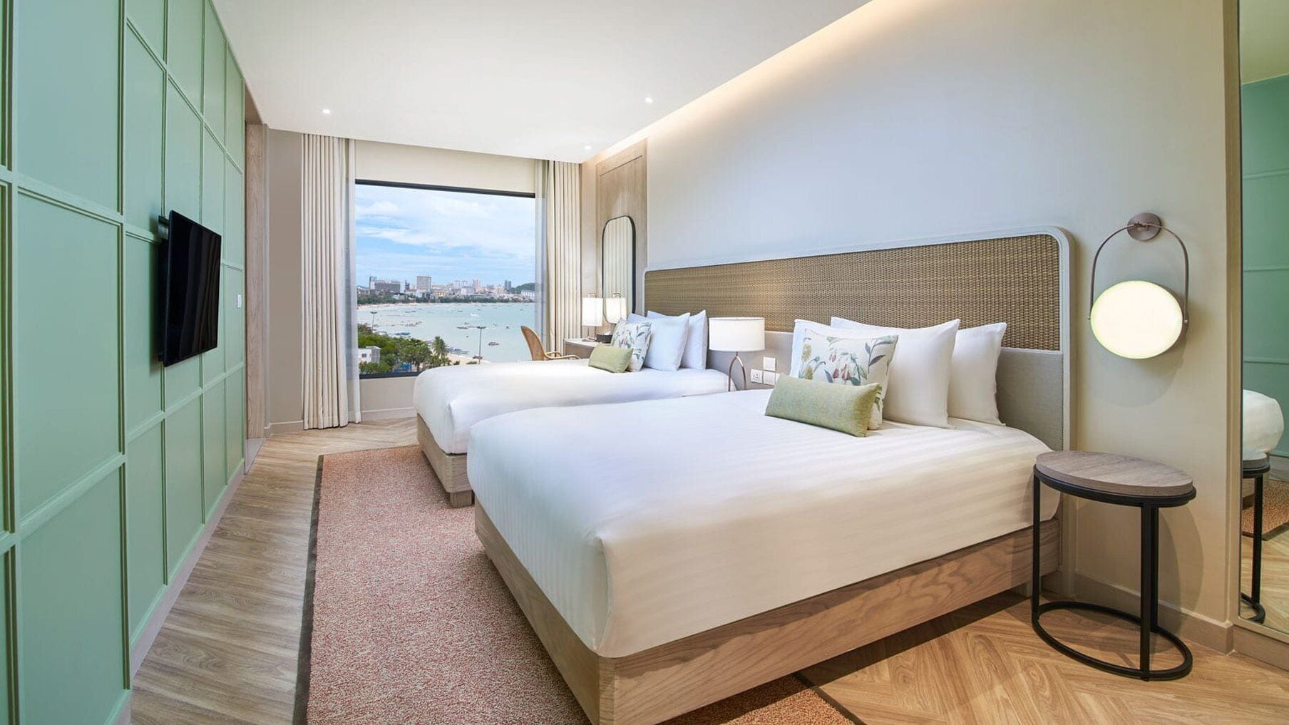 A hotel room with two beds, a large window with a city view, a flat-screen TV, and a round table with a lamp.