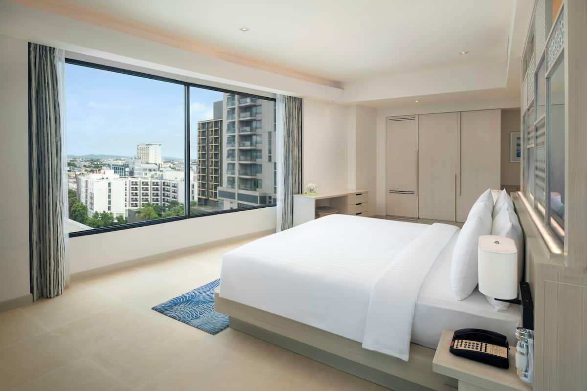 A modern hotel room with a large bed, a bedside telephone, and a view of the city through floor-to-ceiling windows.