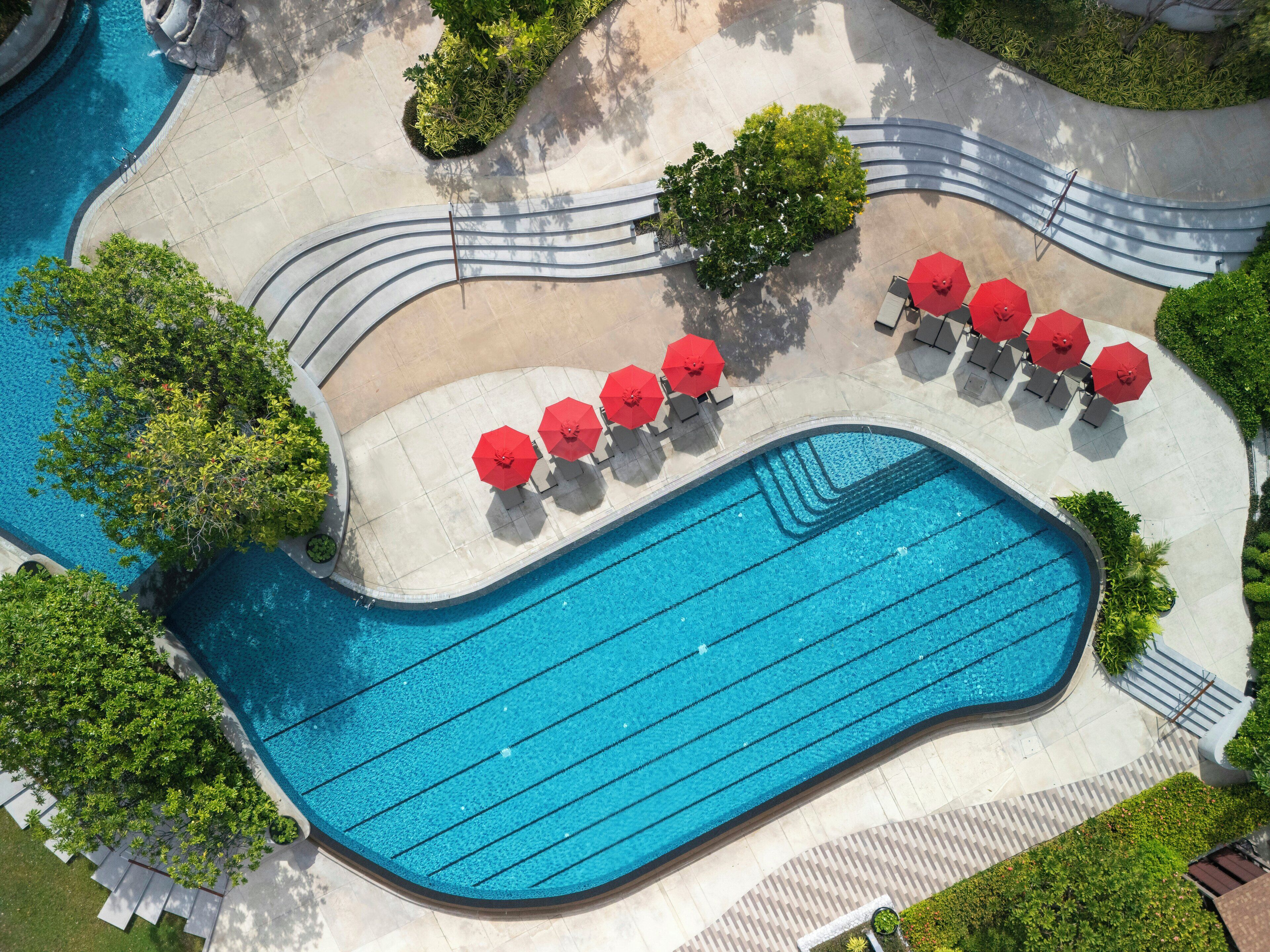 An aerial view of a swimming pool area with a curved pool, red umbrellas, and lounge chairs.