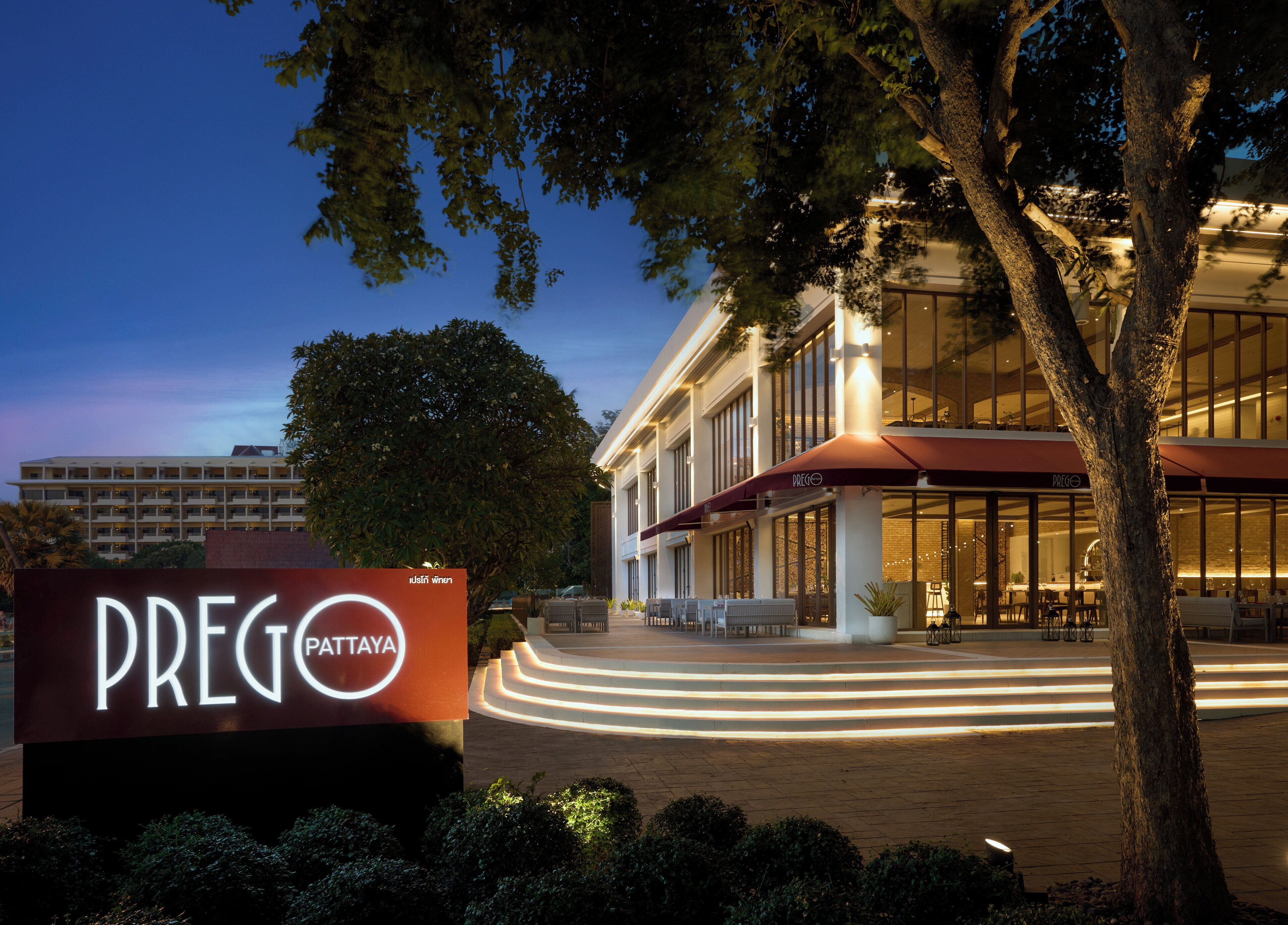 Entrada de un hotel con un cartel que dice "PREGO PATTAYA" y un edificio moderno con fachada de vidrio y zona de estar al aire libre.