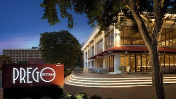 A hotel entrance with a sign reading "PREGO PATTAYA" and a modern building with a glass facade and outdoor seating area.