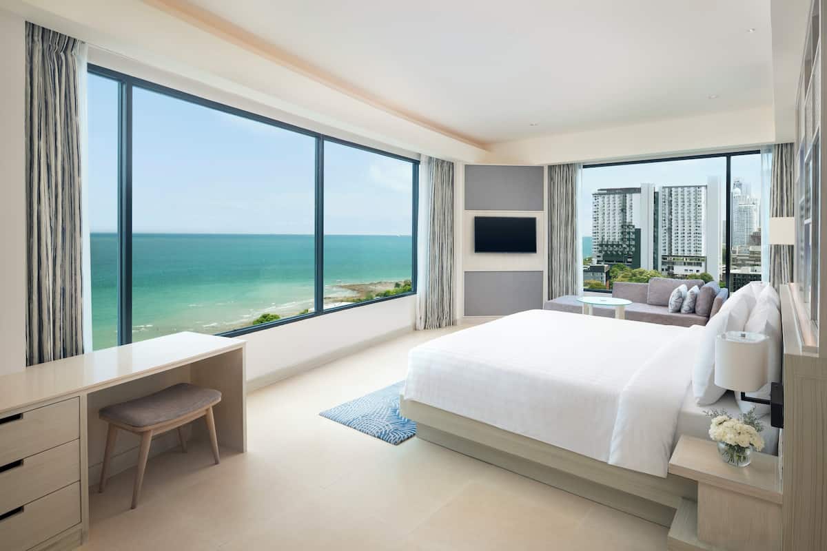 A modern hotel room with a large bed, a desk, and a TV. It has a view of the ocean and tall buildings.
