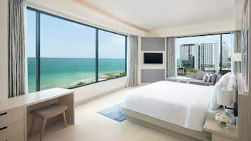 A modern hotel room with a large bed, a desk, and a TV. It has a view of the ocean and tall buildings.