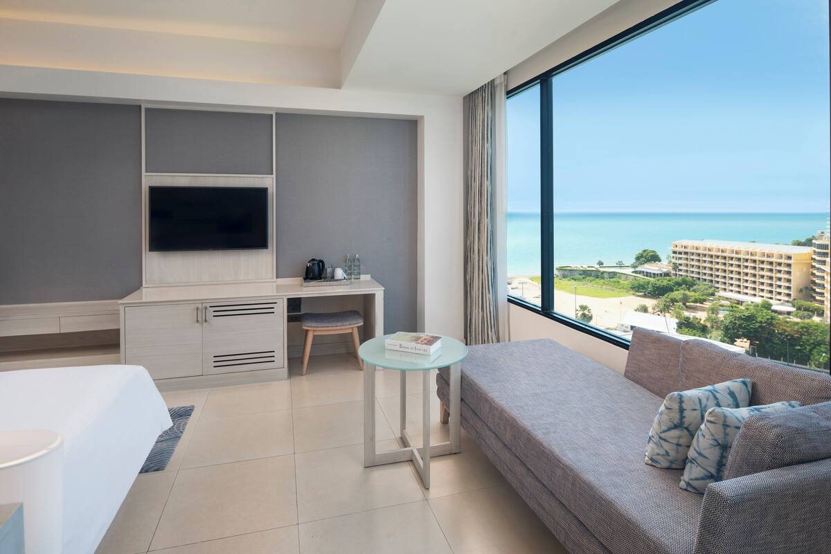 A modern hotel room with a large window offering a view of the ocean and cityscape, a comfortable sofa, a desk with a flat-screen TV, and a bed.