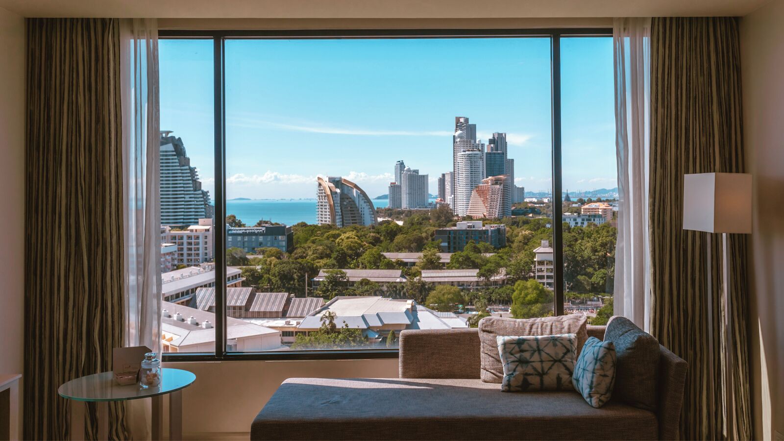 A room with a large window offering a city view, a sofa with cushions, a small table, and a lamp.