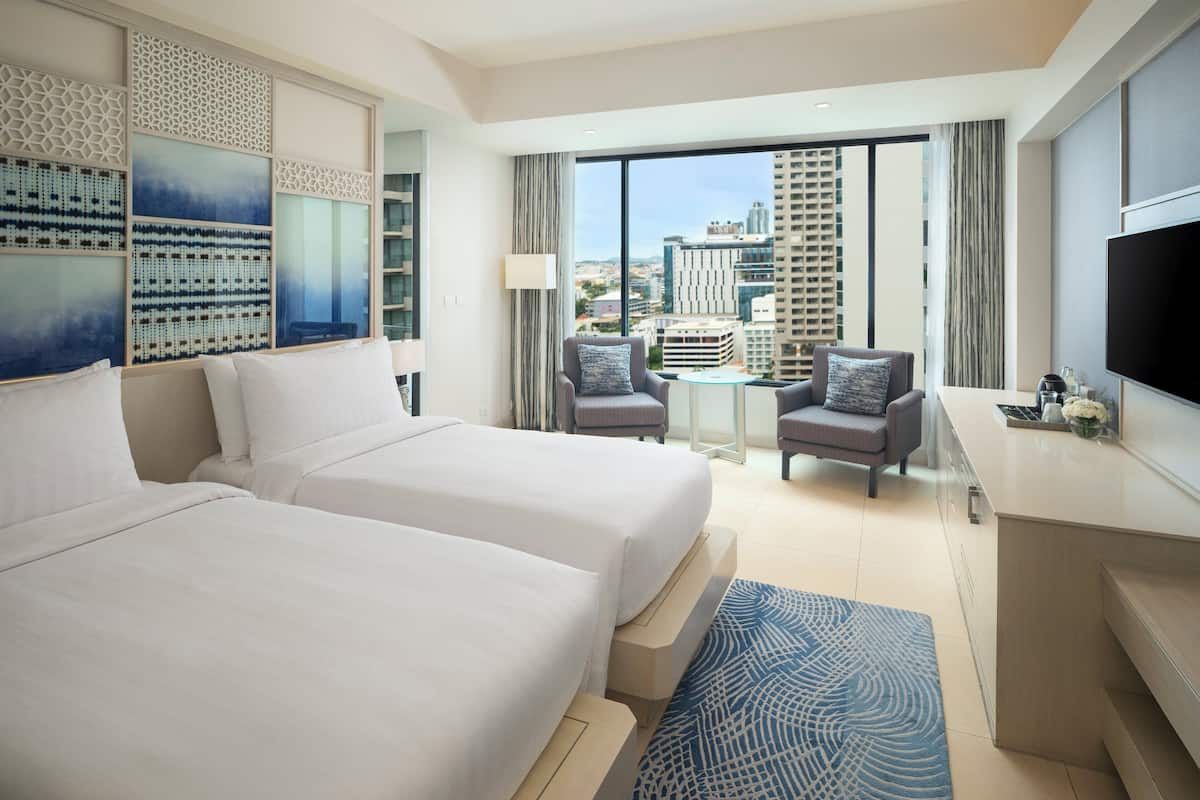 A modern hotel room with two beds, a small dining area, and a large window offering a city view.