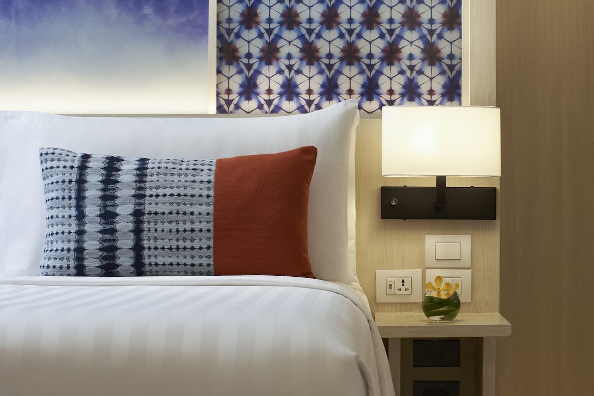 A neatly made bed with white bedding and patterned pillows, a bedside table with a lamp, and a wall with a geometric patterned design.