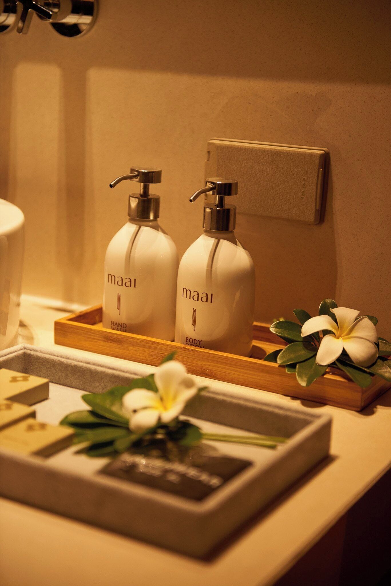 Un tocador de baño con dos botellas de jabón líquido 'maai' para manos y cuerpo, una bandeja de madera y un pequeño florero con una flor blanca.