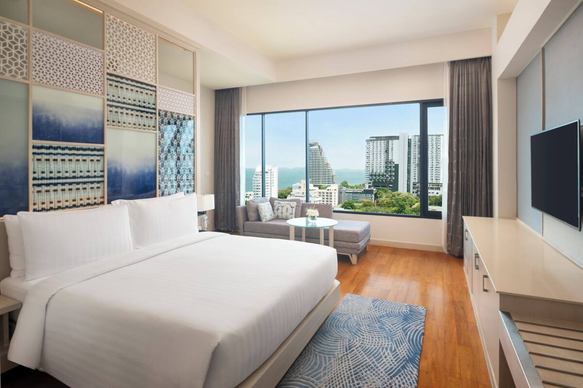 A modern hotel room with a large bed, a sofa, a small table, and a TV. The room has large windows with a city view.