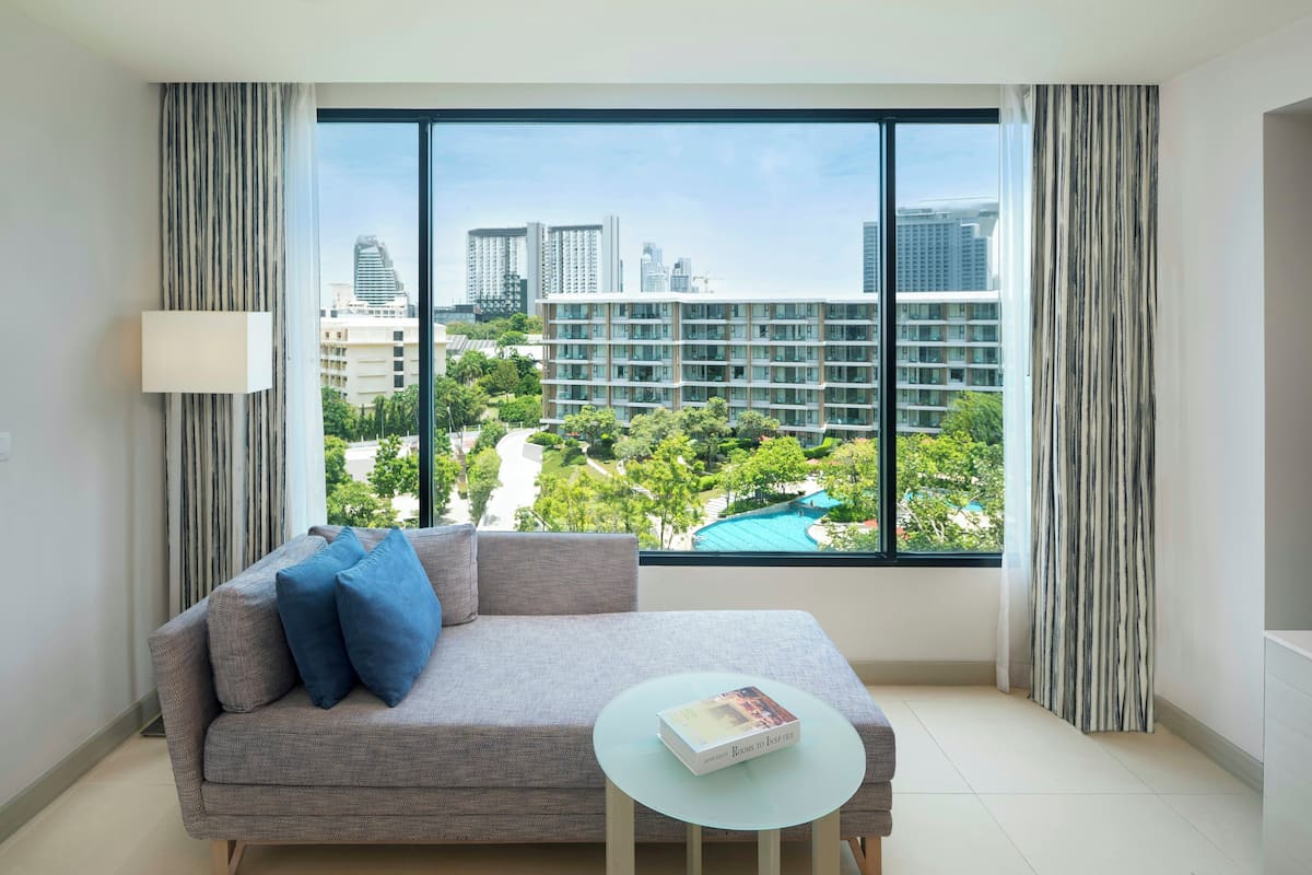 A modern living room with a grey sofa, a small round table, and a book. A large window offers a view of a cityscape and a swimming pool.