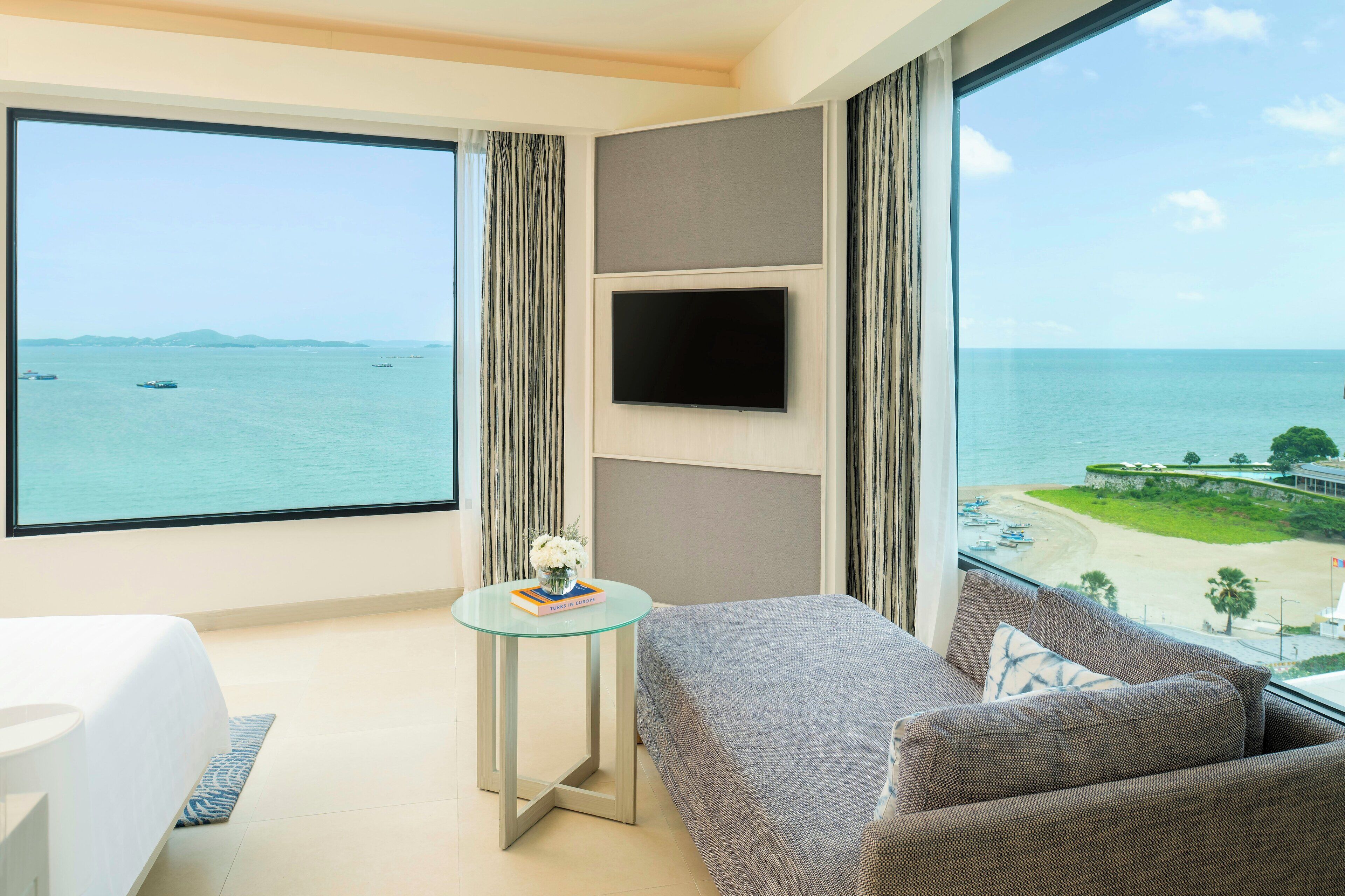 A hotel room with a large window, a bed, a sofa, a small table with a book and flowers, and a flat-screen TV mounted on the wall.