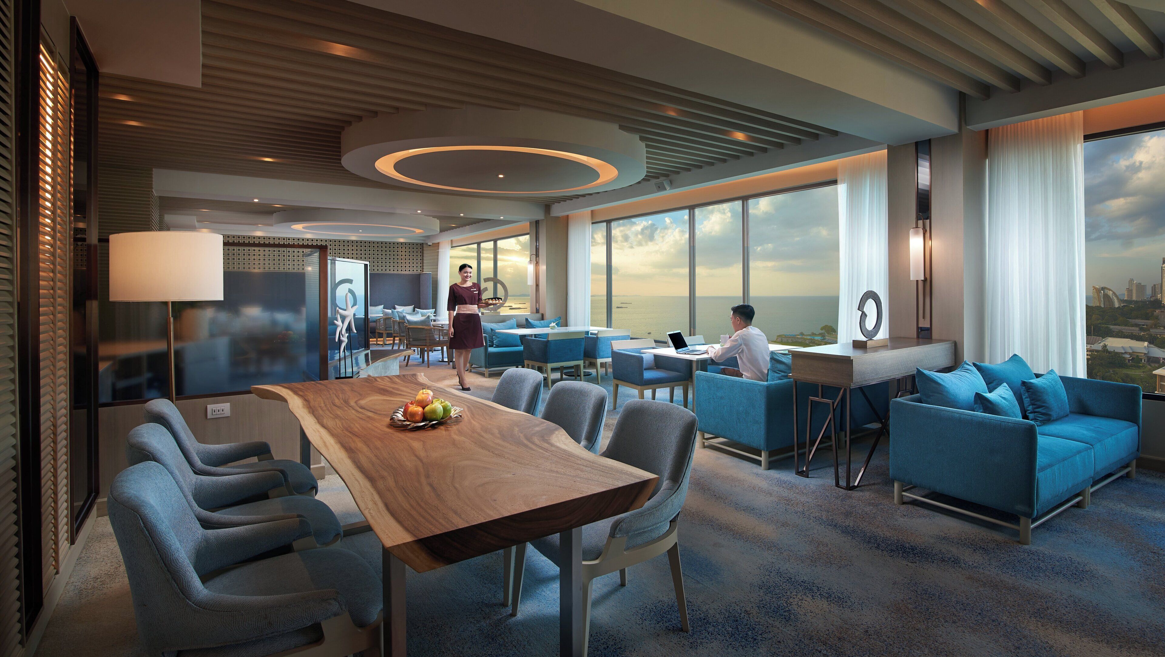 A modern hotel lobby with a wooden reception desk, blue seating, and large windows offering a view of the city skyline.