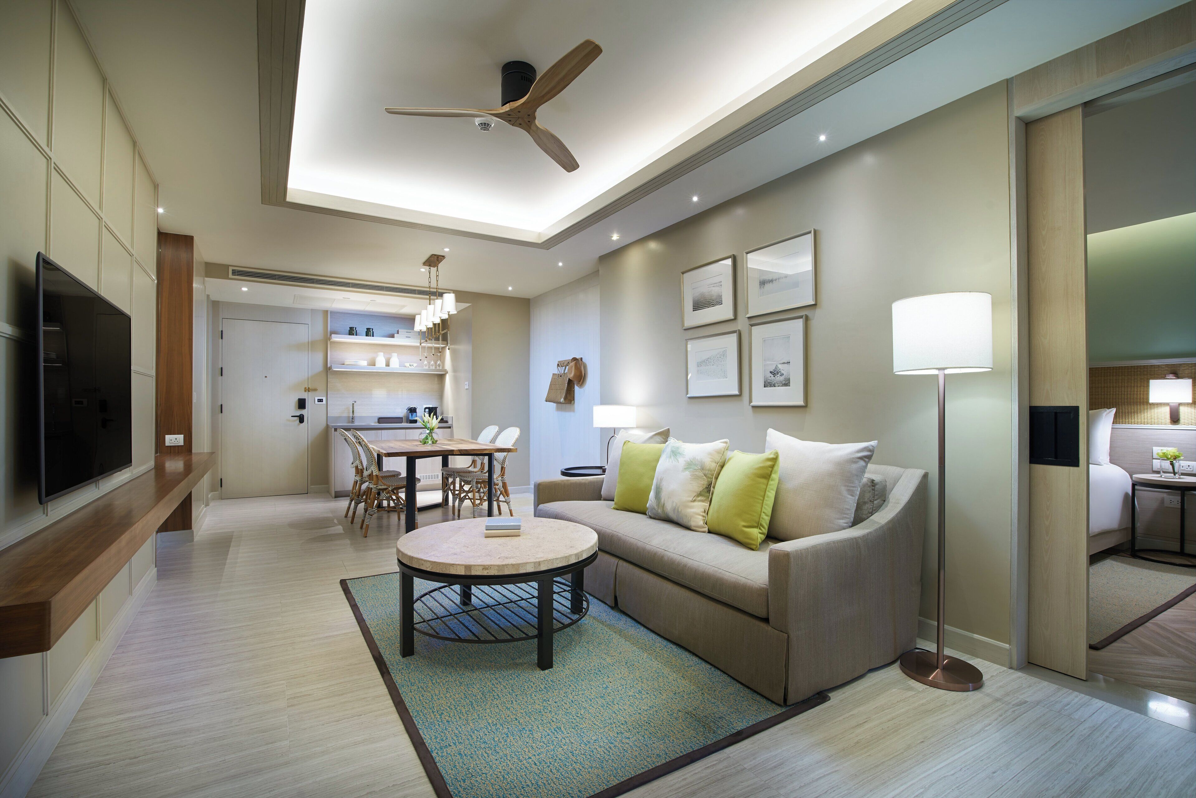 Un salon moderne avec un canapé, une table basse et un ventilateur de plafond. On y trouve également un coin repas avec des chaises et une table, ainsi qu’une cuisine en arrière-plan.