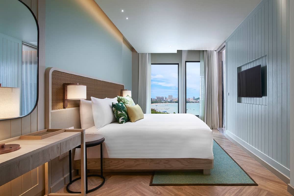 A hotel room with a large bed, a desk with a lamp, a TV mounted on the wall, and a view of the city through large windows.