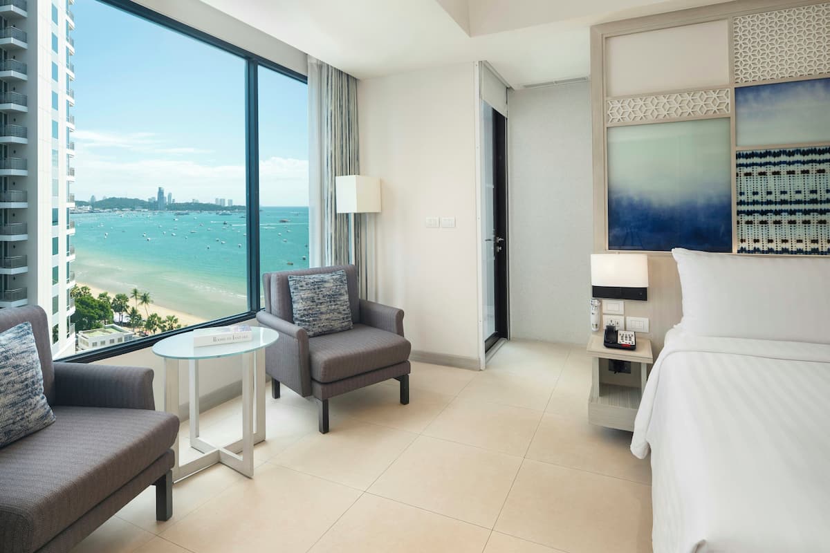 A hotel room with a large window offering a view of the beach and ocean. There is a bed, a chair, a small table, and a nightstand with a phone.