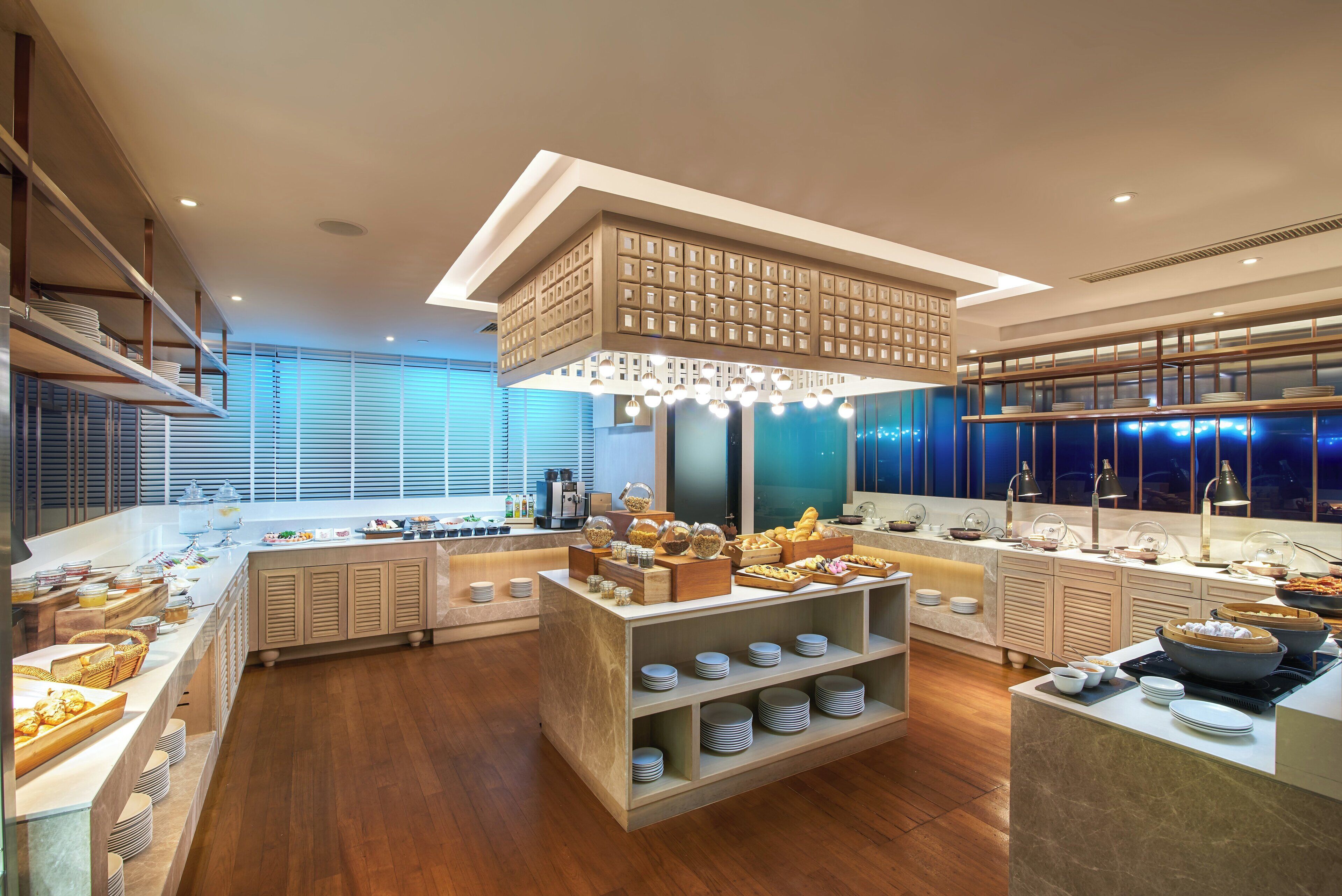 Un área de desayuno de hotel moderna con diversas opciones de comida, una estación central de servicio y un amplio ventanal que proporciona luz natural.