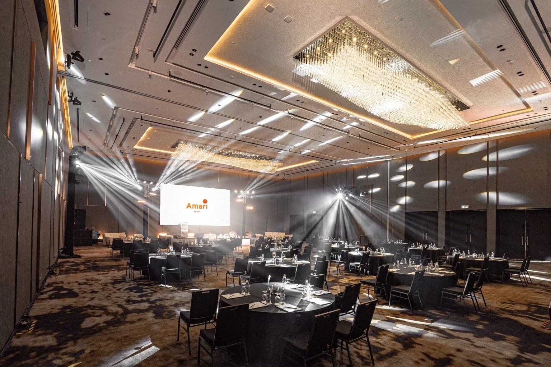 A large conference room with tables and chairs, a large screen displaying 'Amari', and a chandelier.